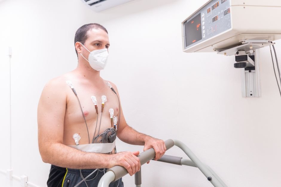 Hombre en examen médico con electrodos y máscara facial realizando prueba de estrés cardíaco en la cinta de correr.